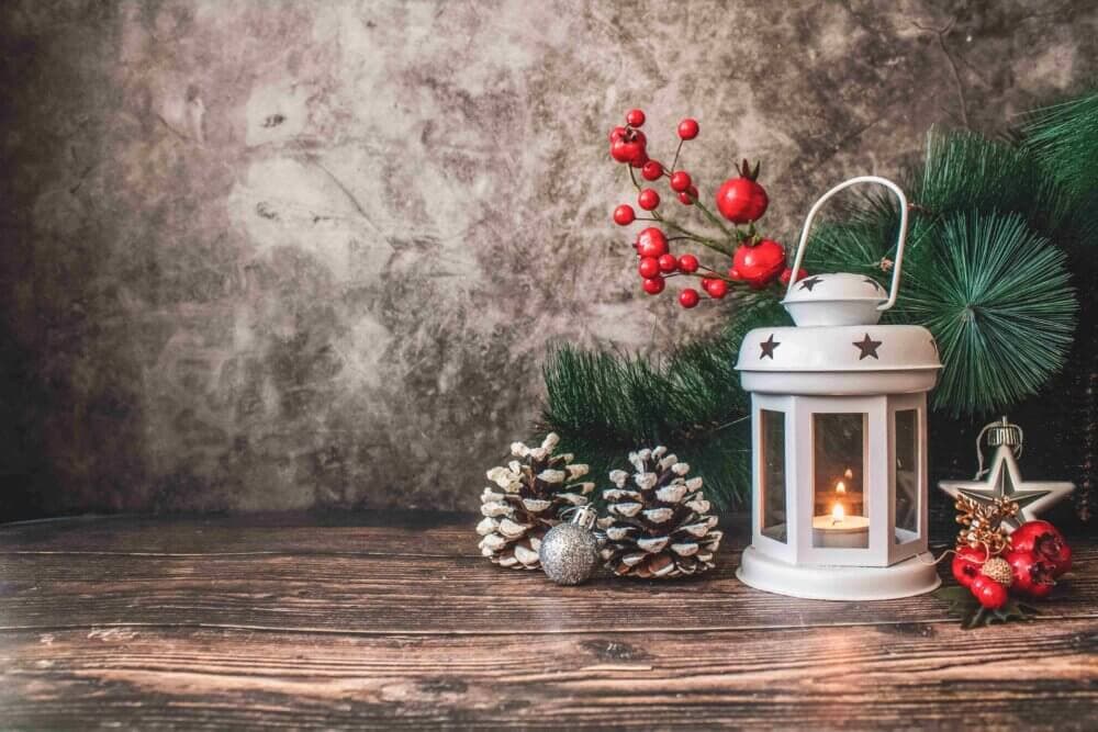 A lit lantern with festive decorations, including pinecones, red berries, and green pine branches on a wooden surface. - Home Instead