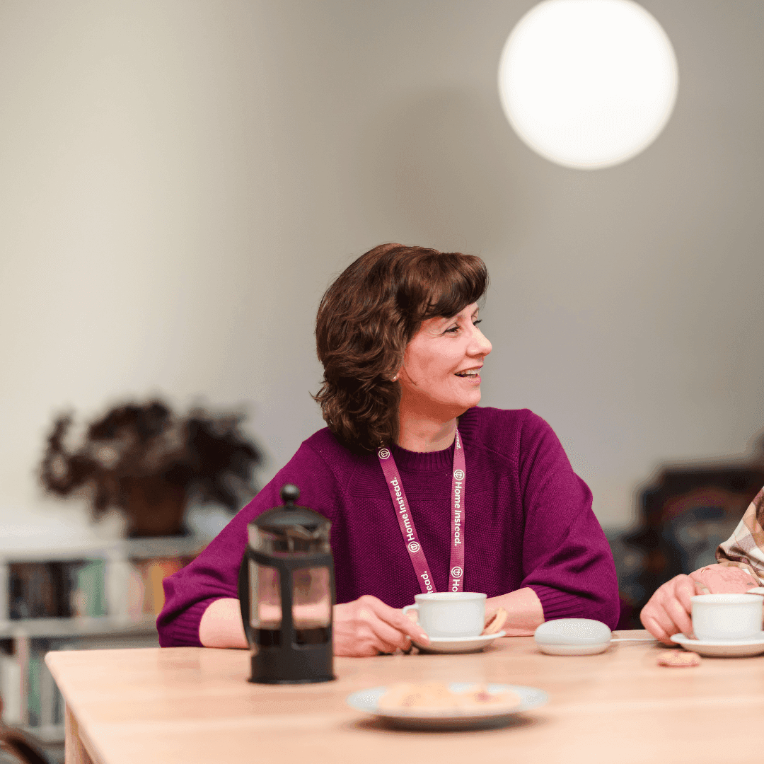 Person smiling at a table with coffee, pastries, a French press, and a white device. Shelves and plants in the background. - Home Instead