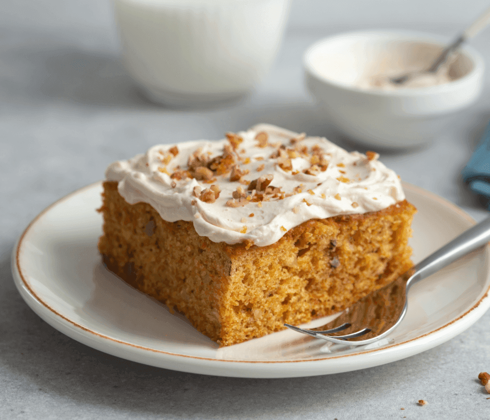 A slice of carrot cake with frosting and chopped nuts on a plate with a fork, milk, and a bowl in the background. - Home Instead