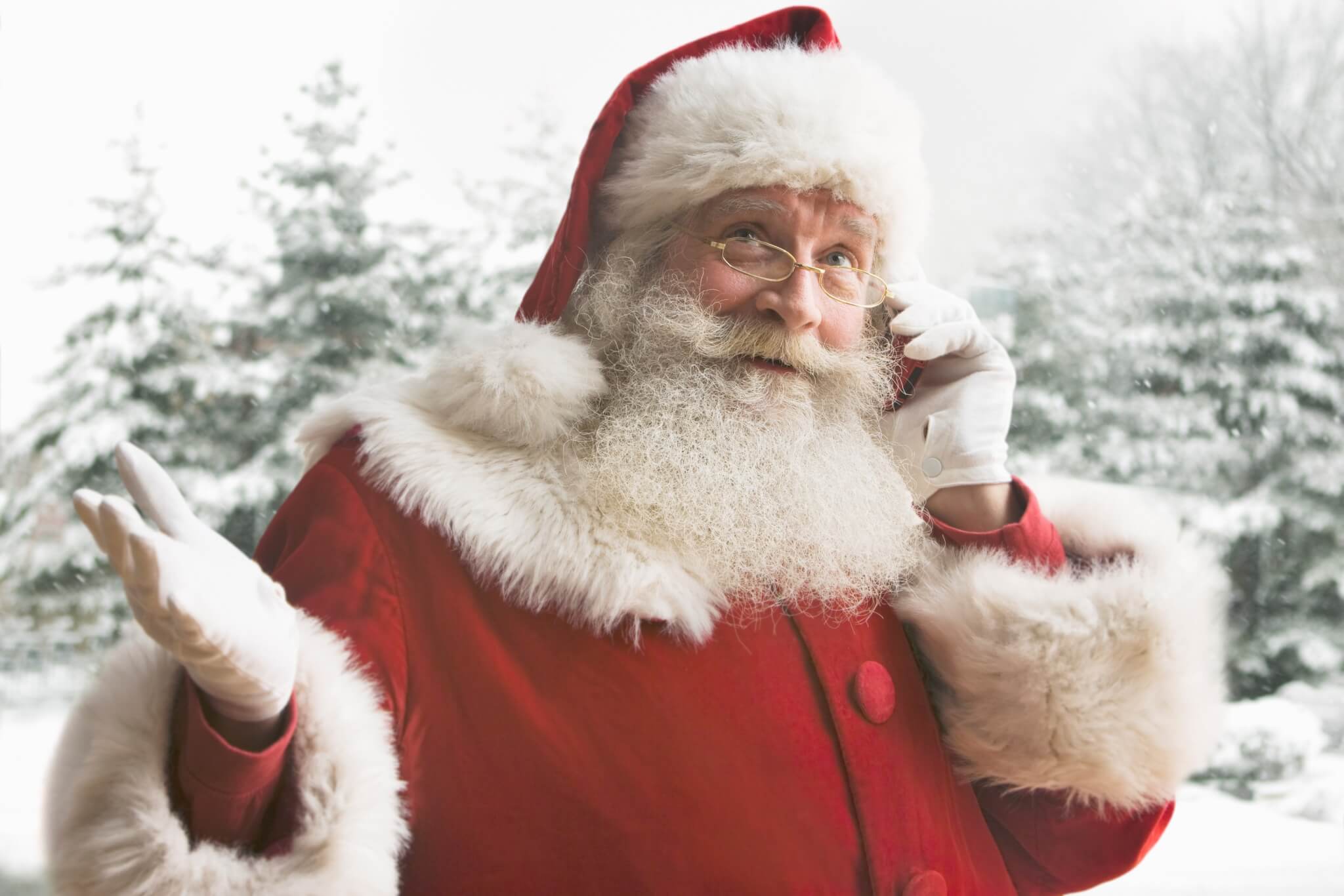 Jolly Santa Claus in red suit and glasses, talking on a cell phone, with snowy trees in the background. - Home Instead