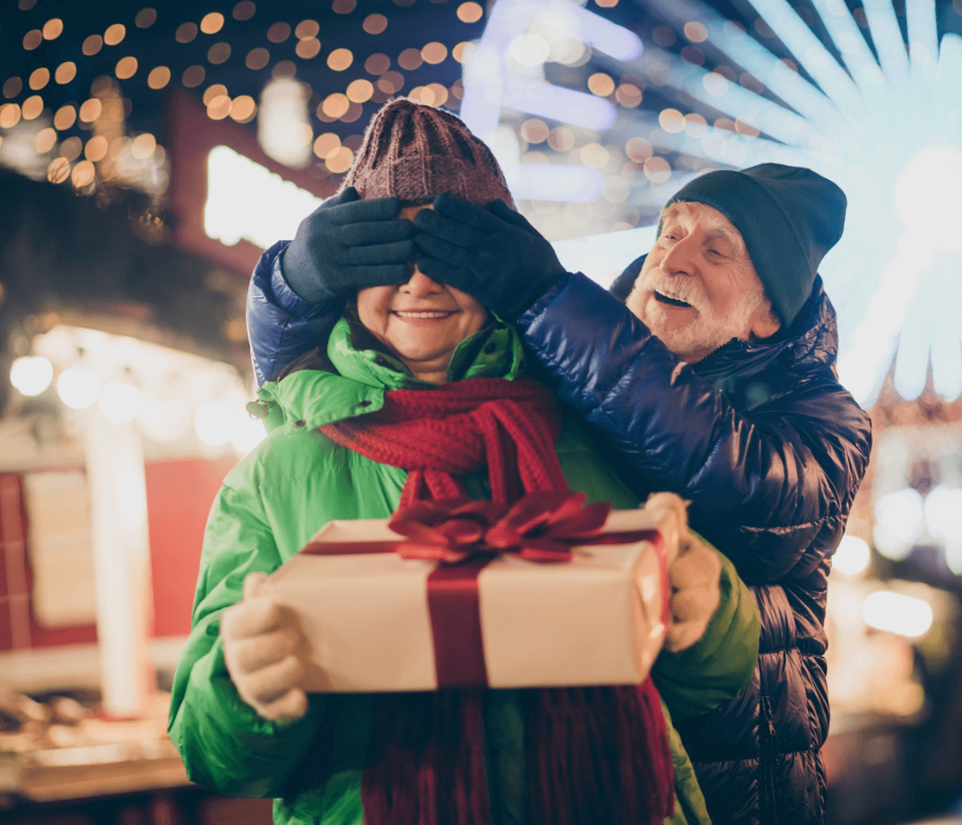 An elderly man surprises a woman with a gift, covering her eyes at a festive outdoor event, with lights in the background. - Home Instead