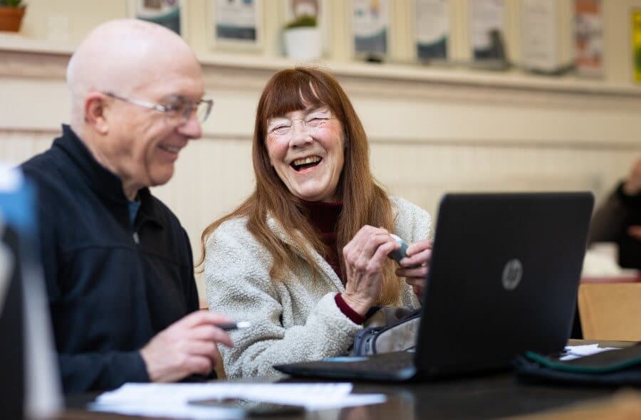 Two older adults smiling while using a laptop together in a cozy room. - Home Instead