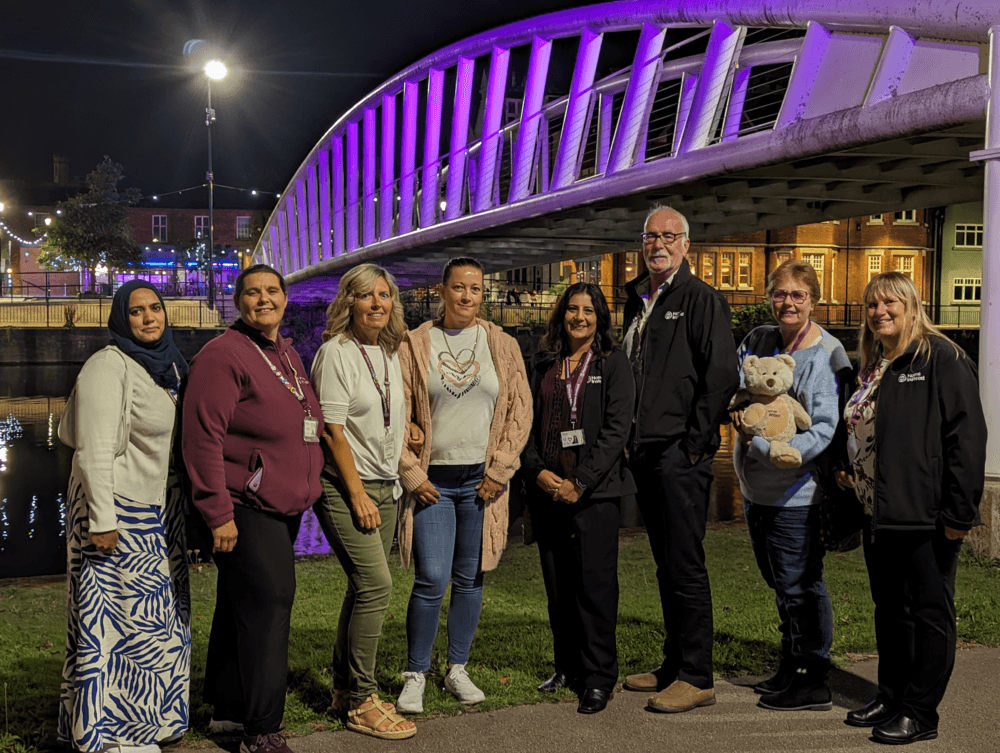 A group of people standing together in front of a bridge illuminated with purple lights at night. - Home Instead