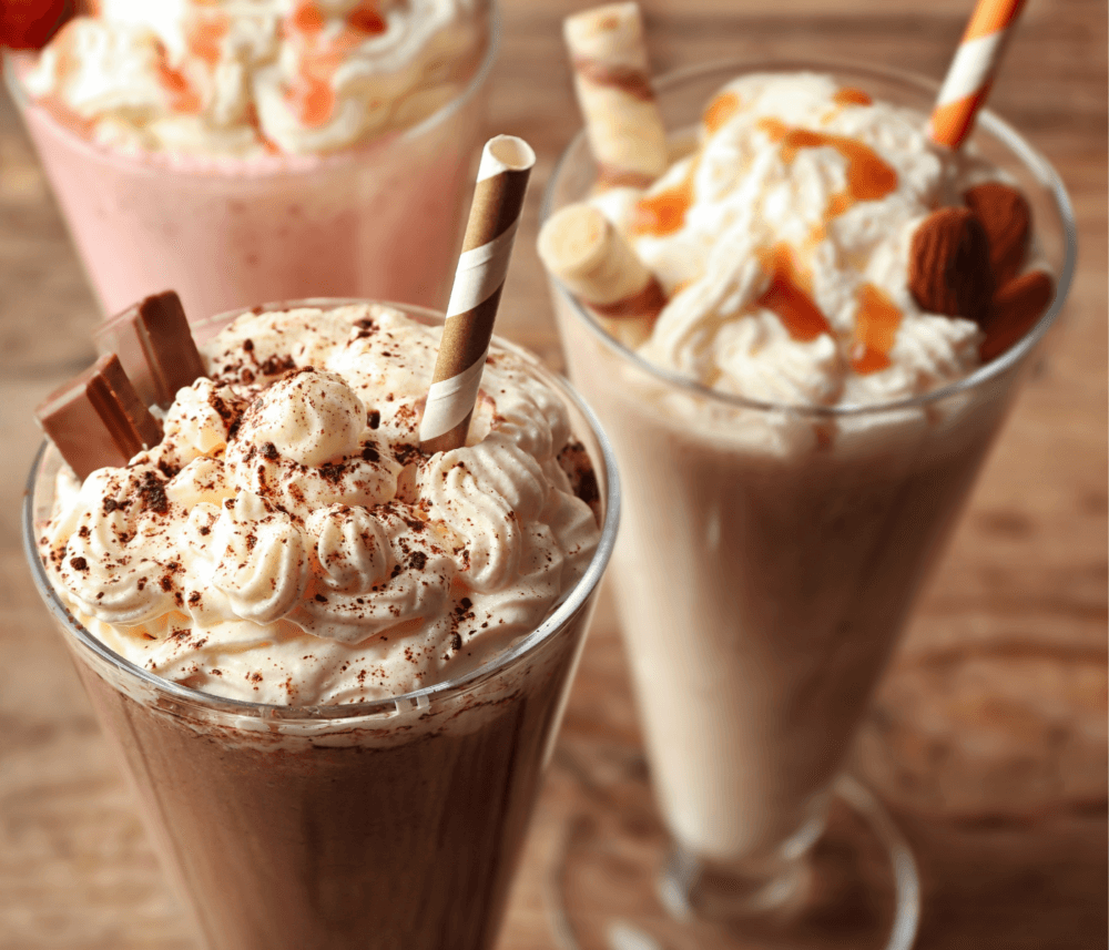 Three milkshakes topped with whipped cream, chocolate shavings, and caramel sauce on a wooden table. - Home Instead