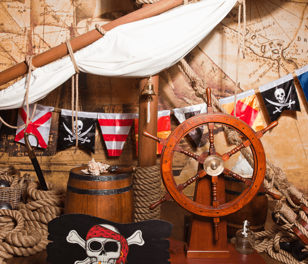 A pirate-themed decoration with flags, a ship wheel, a barrel, nautical ropes, and a pirate skull. - Home Instead