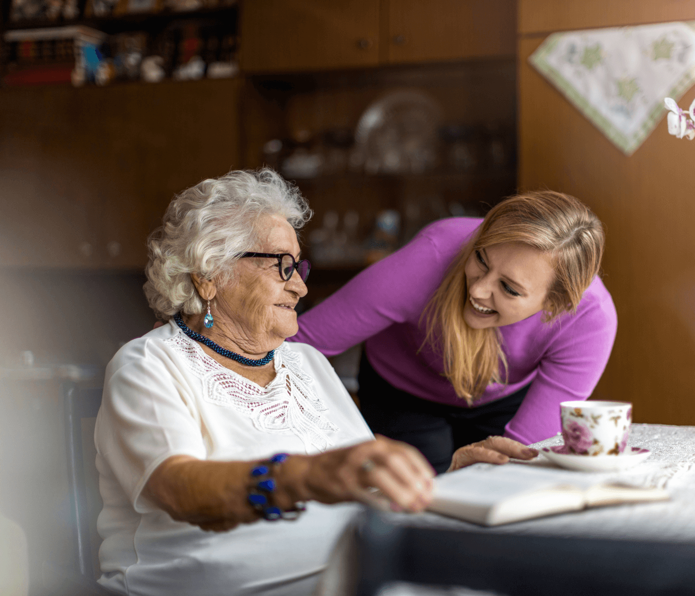 Elderly woman with glasses and young woman smiling at each other in front of table with a book and teacup. - Home Instead