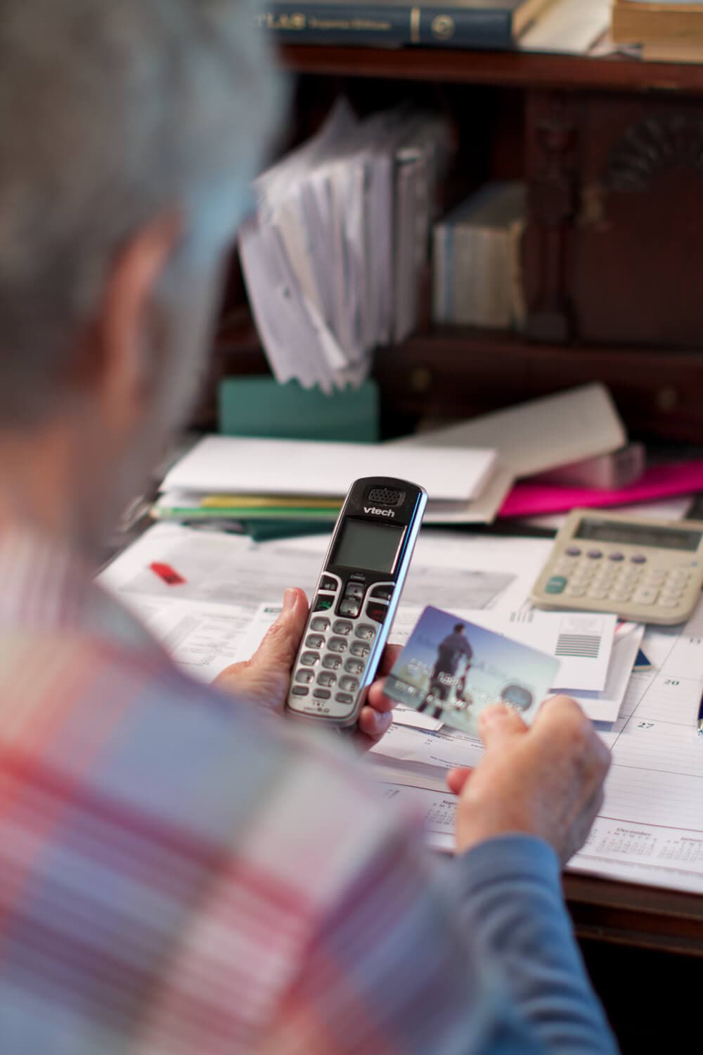 Person holding a landline phone and a photograph, sitting at a cluttered desk with papers, calculator, and books. - Home Instead