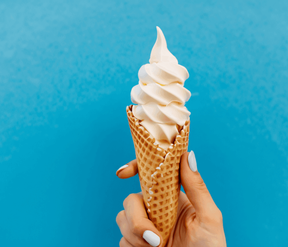 A hand holding a vanilla soft serve ice cream cone against a bright blue background. - Home Instead