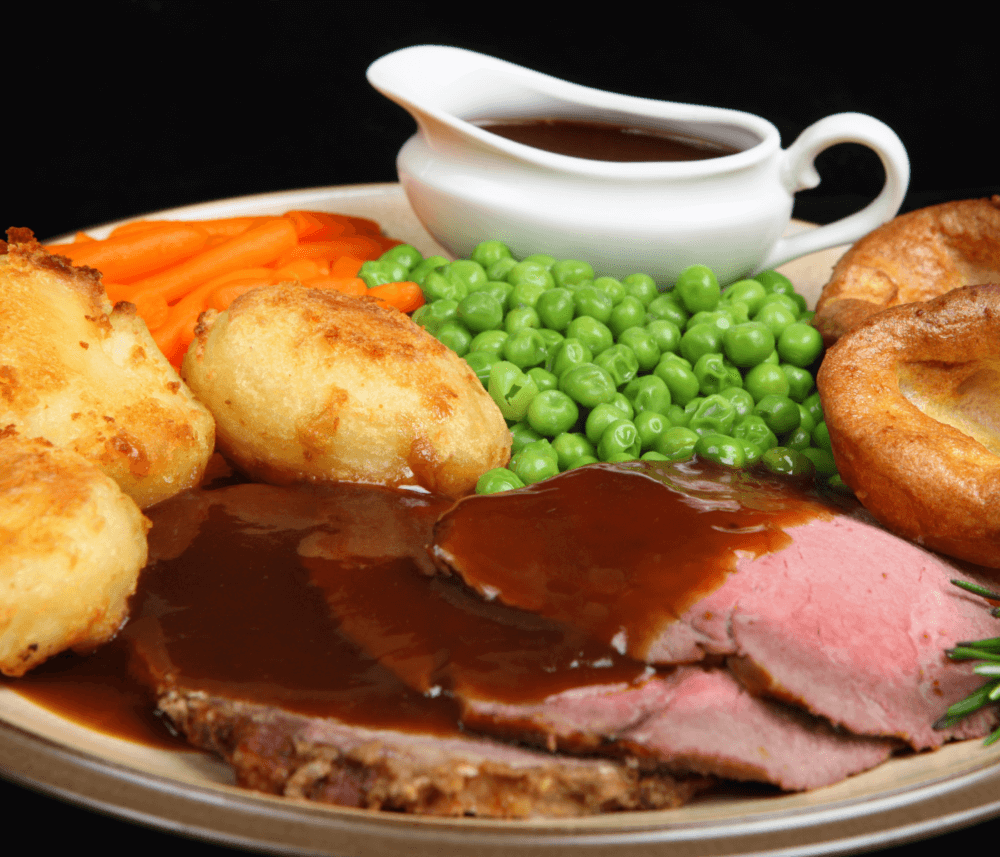 A plate of roast beef with gravy, peas, carrots, Yorkshire pudding, and roast potatoes, with a gravy boat in the background. - Home Instead