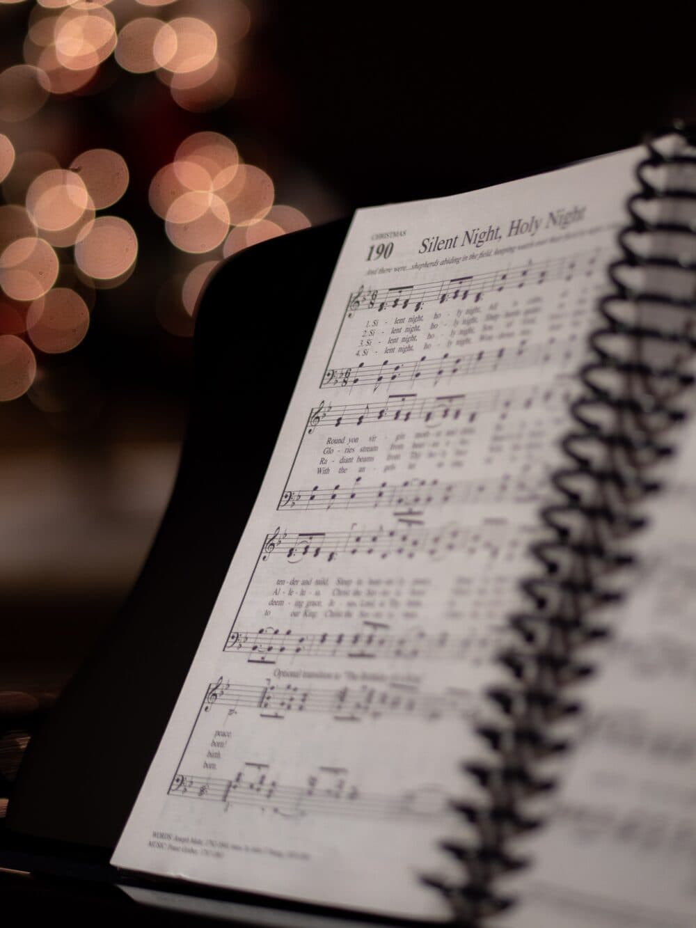 Close-up of an open hymnbook with "Silent Night, Holy Night" sheet music, and bokeh Christmas lights in the background. - Home Instead