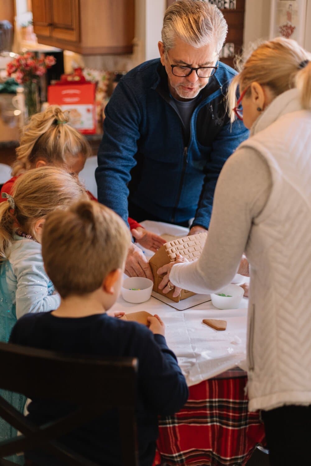 An older man and children gather around a table to decorate a gingerbread house, assisted by a woman in a white vest. - Home Instead