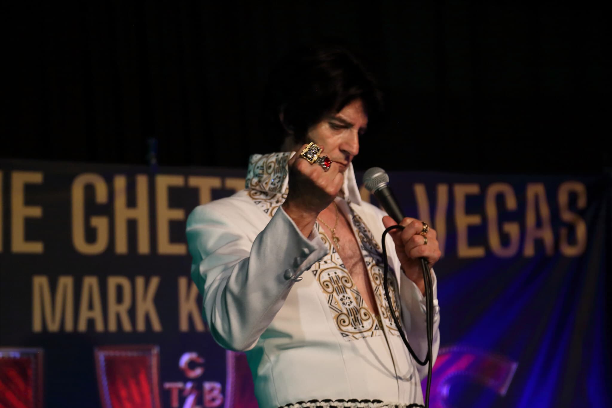 A performer in a white jumpsuit sings into a microphone on stage, with a sign reading "Vegas" in the background. - Home Instead