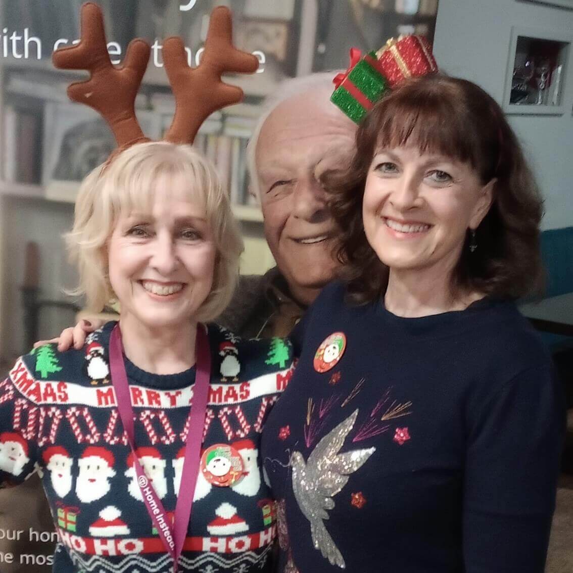 Two women dressed in festive holiday attire are smiling, one wearing reindeer antlers and the other a small Christmas hat. - Home Instead