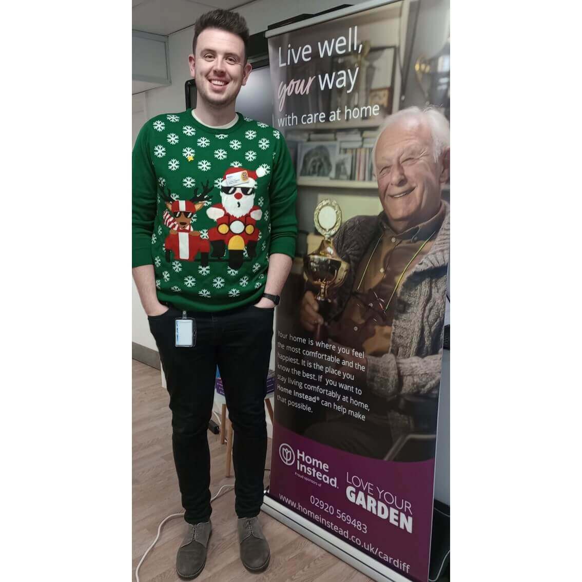 Man in green Christmas sweater stands next to a banner promoting home care for the elderly by Home Instead. - Home Instead