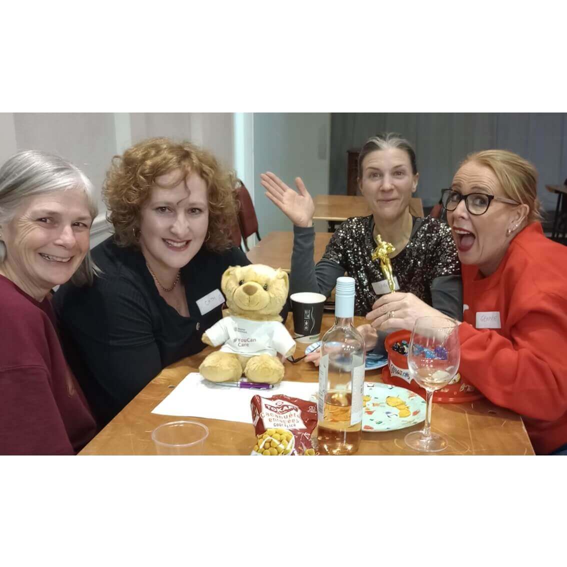 Four women smiling and sitting around a table with a teddy bear, wine bottle, and snacks. One holds a trophy. - Home Instead
