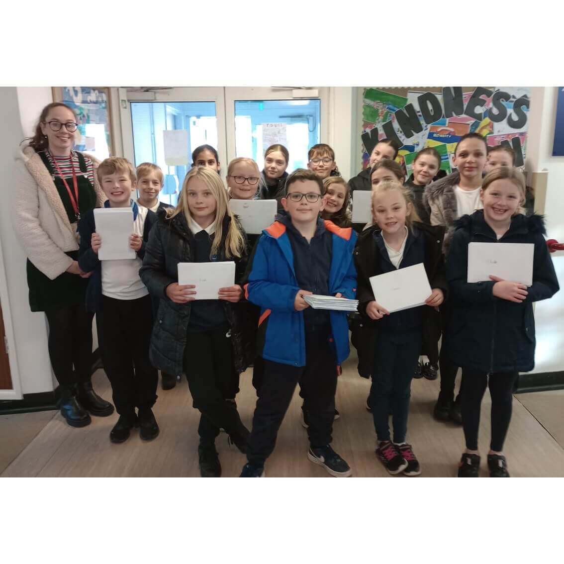 A group of school children and a teacher smile and hold certificates in a hallway near a poster that reads "KINDNESS. - Home Instead