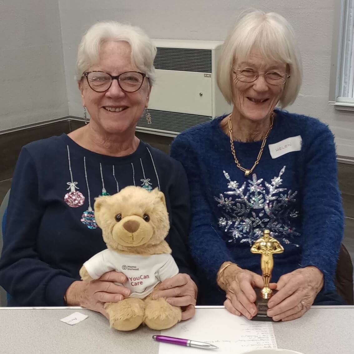 Two older women smiling, one holding a teddy bear, and the other holding a small trophy, seated indoors. - Home Instead