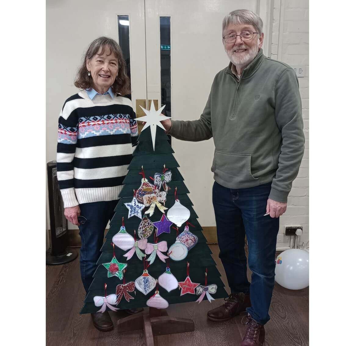 Two smiling people stand beside a small decorated Christmas tree indoors. A white balloon is on the floor nearby. - Home Instead