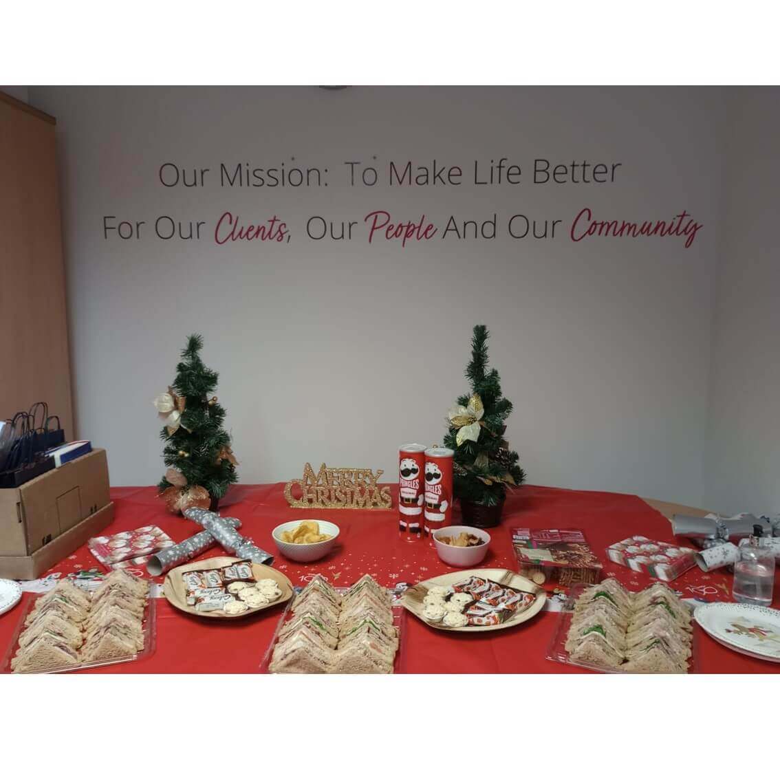 A festive table with sandwiches and holiday treats, and a wall with the mission statement "To Make Life Better. - Home Instead