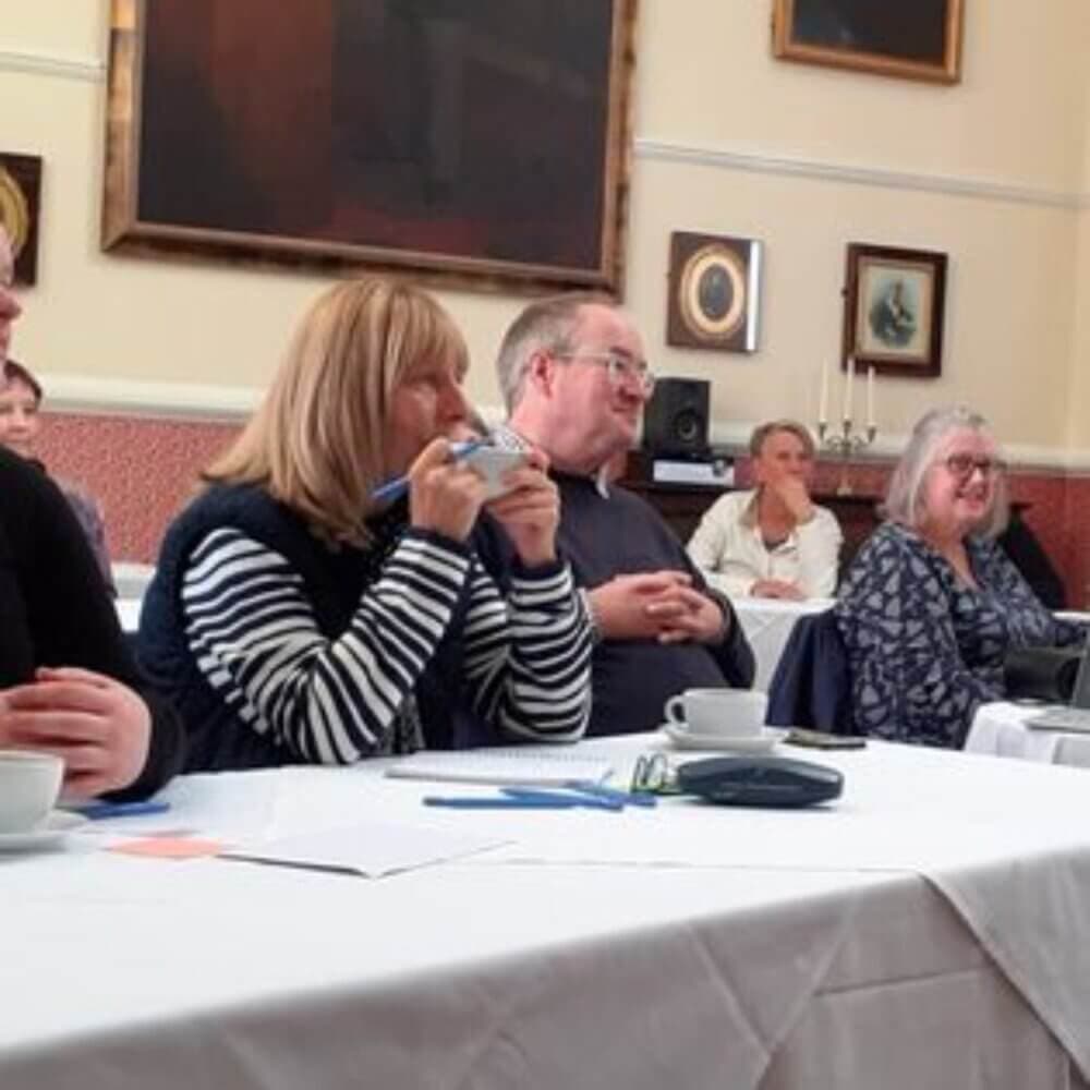 People seated at tables in a meeting room, some taking notes and engaged in a discussion. - Home Instead