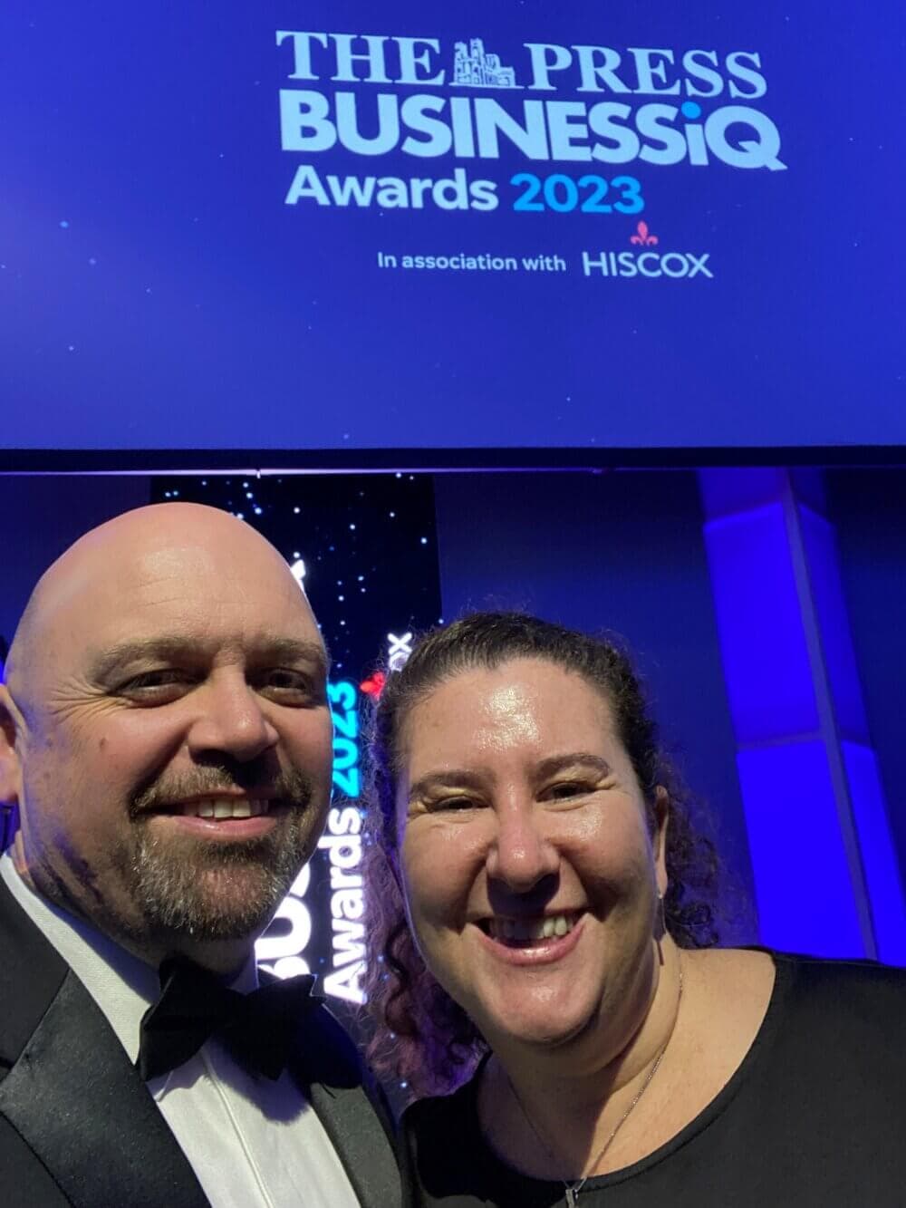 Two people smiling in front of a The Press Business IQ Awards 2023 sign. - Home Instead