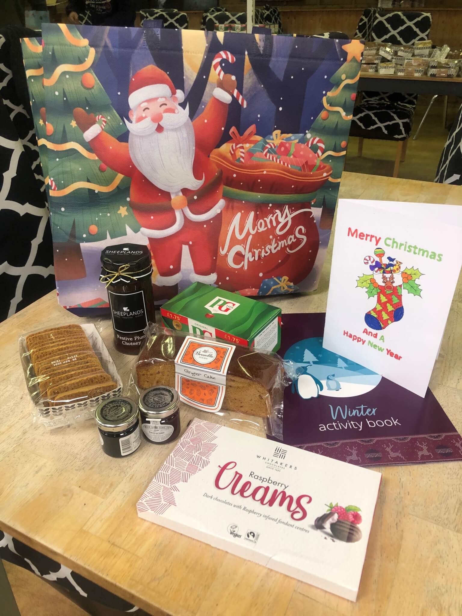 Christmas gift bag with a Santa design, containing treats like biscuits, tea, jams, a card, and a winter activity book. - Home Instead