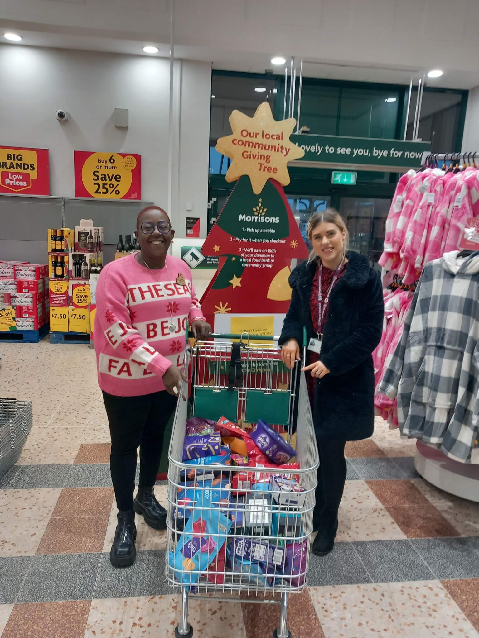 Two smiling people with a shopping cart full of items in front of a Christmas donation tree in a store. - Home Instead