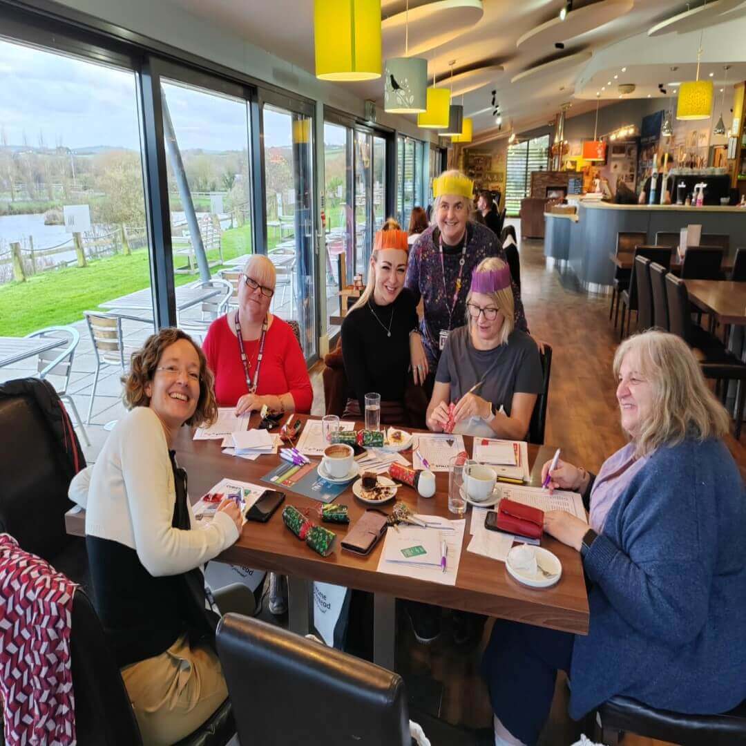 Group of six women sitting around a table in a cafe, smiling, with papers, markers, and drinks on the table. - Home Instead
