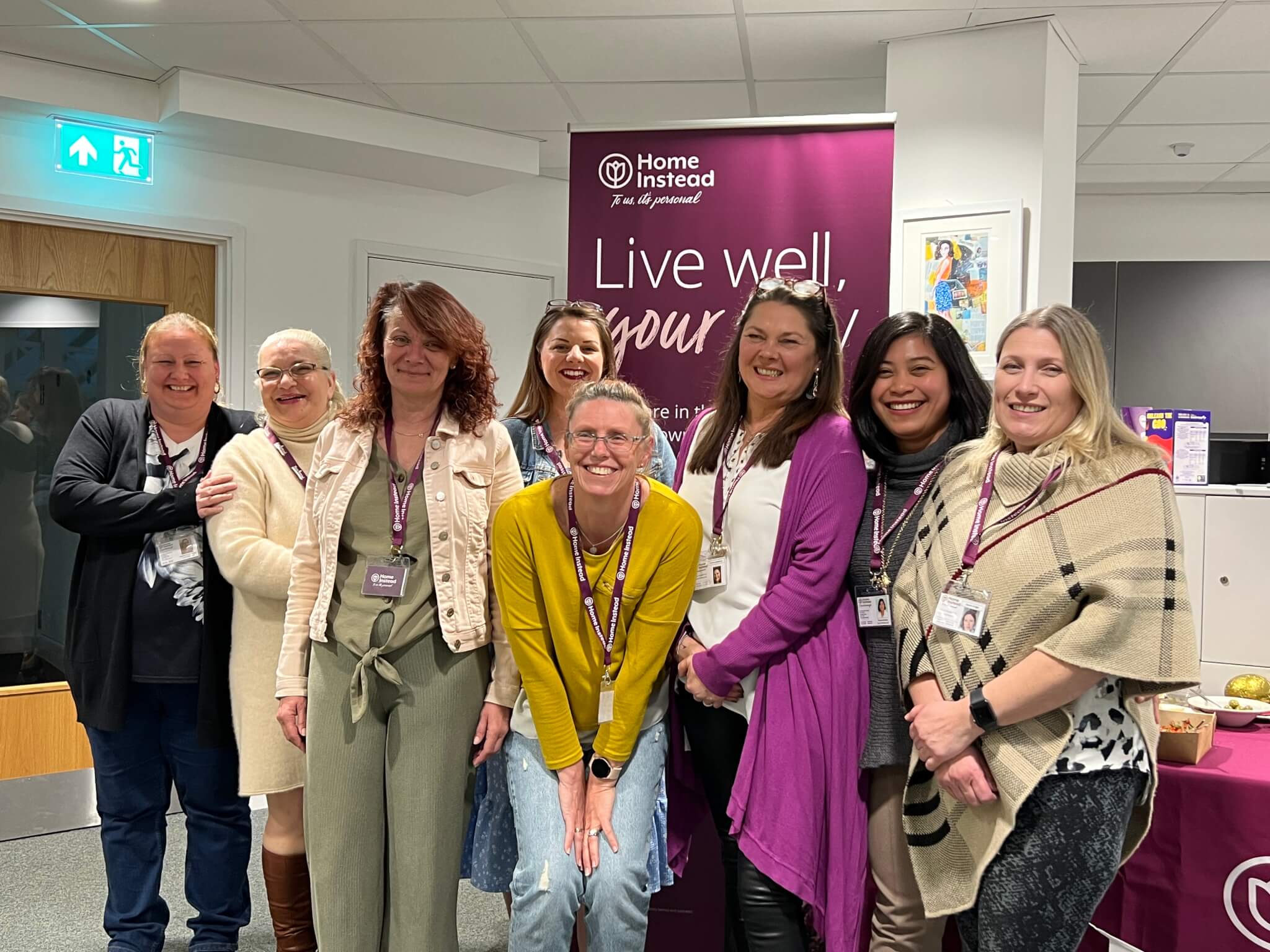 A group of eight smiling women standing together in an office with a "Home Instead" banner in the background. - Home Instead