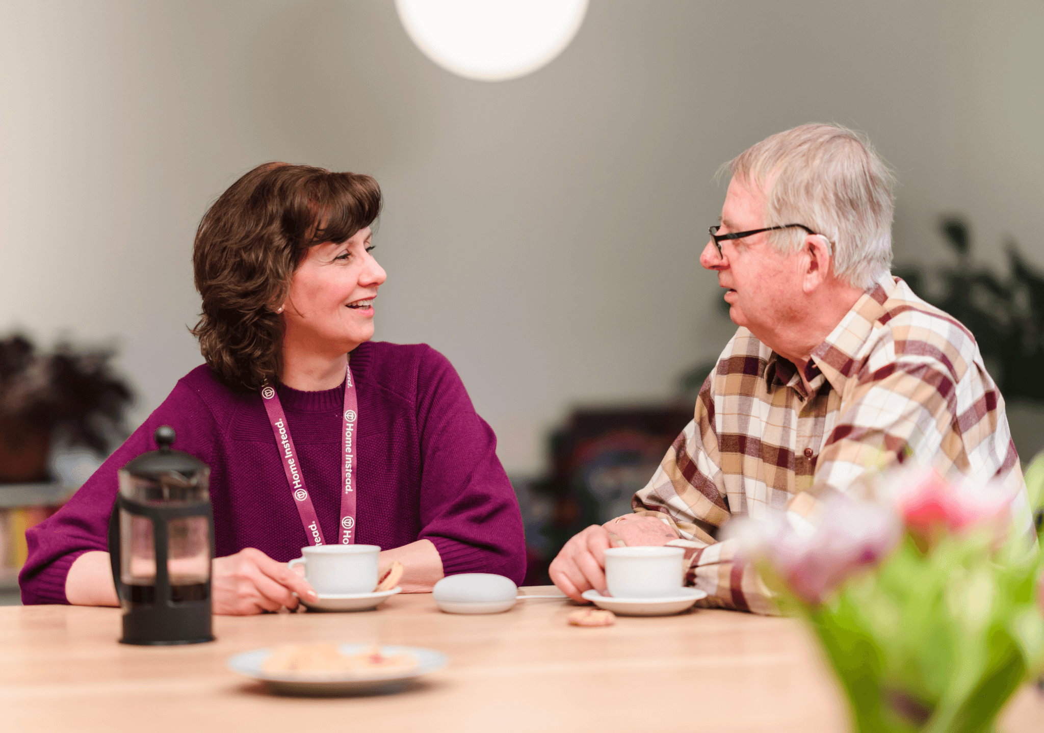 Two elderly people sitting at a table, drinking tea and smiling at each other. - Home Instead
