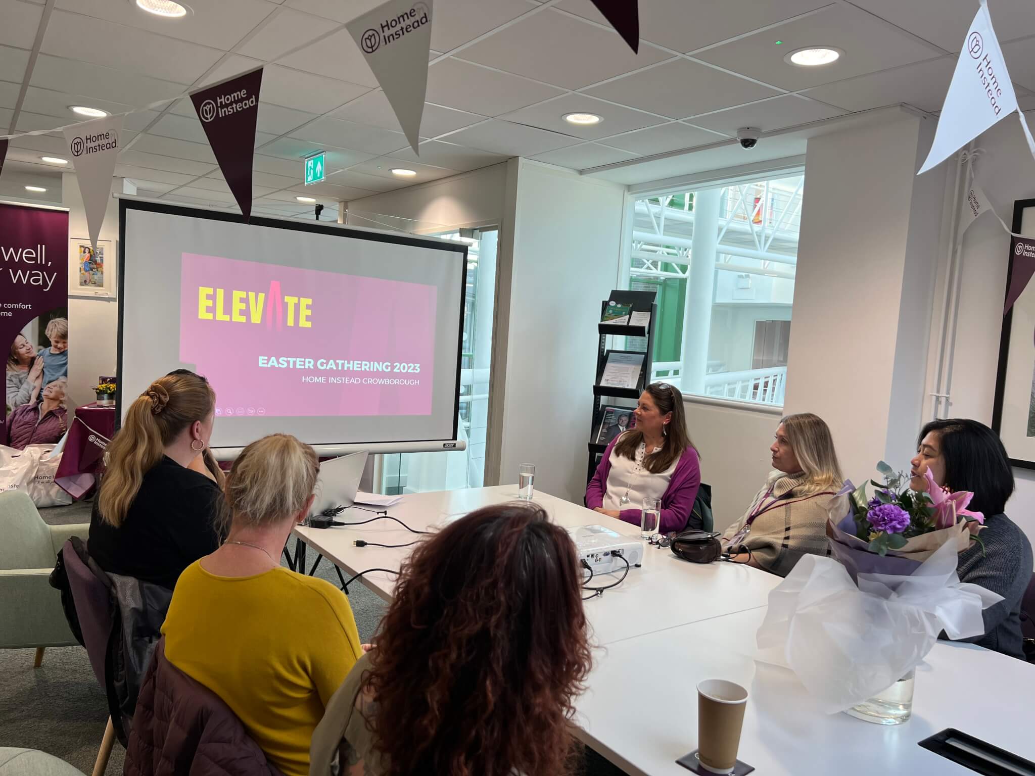 People sitting around a table in an office watching a presentation titled "ELEVATE: Easter Gathering 2023". - Home Instead