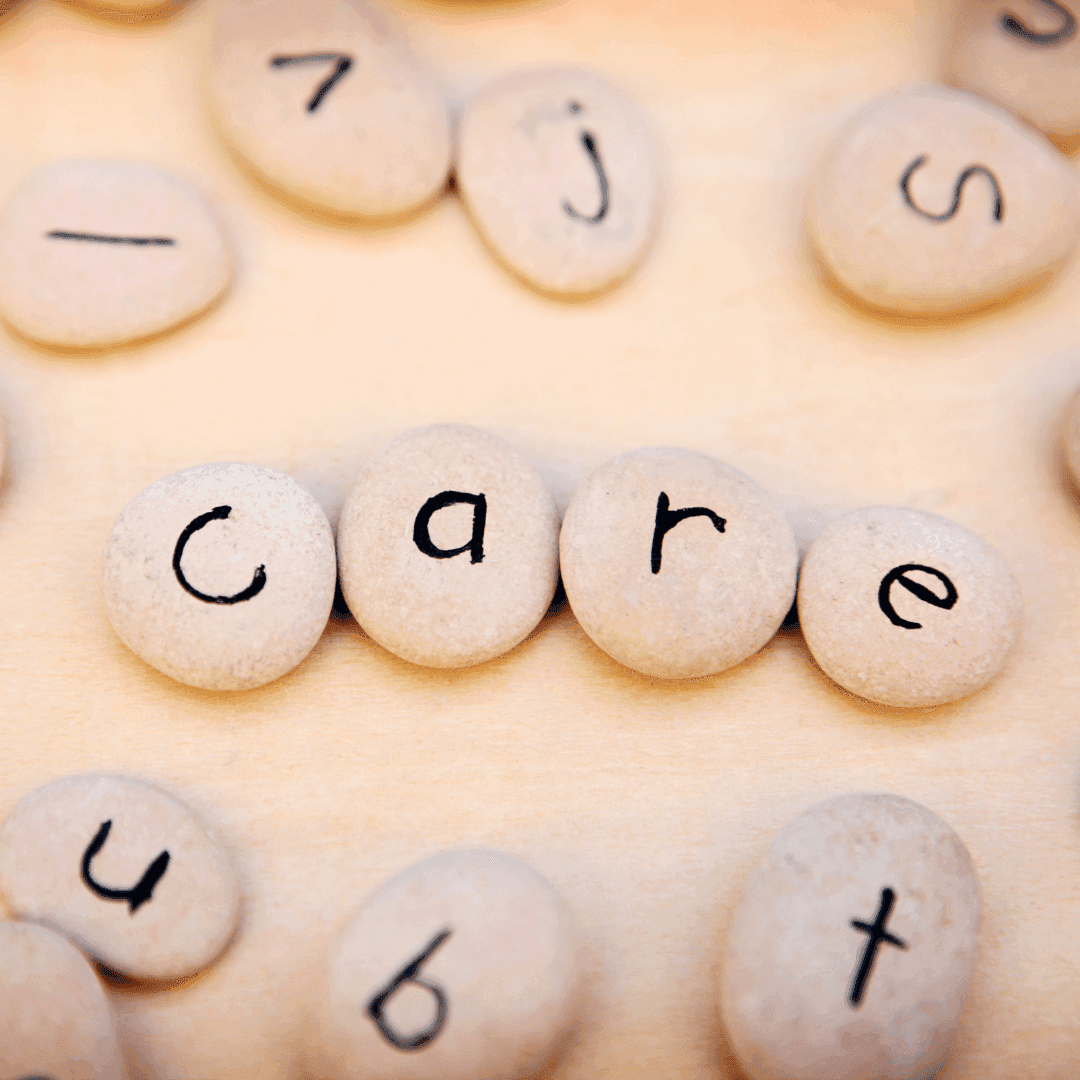 Pebbles with letters written on them form the word "care" on a light surface, surrounded by other lettered pebbles. - Home Instead
