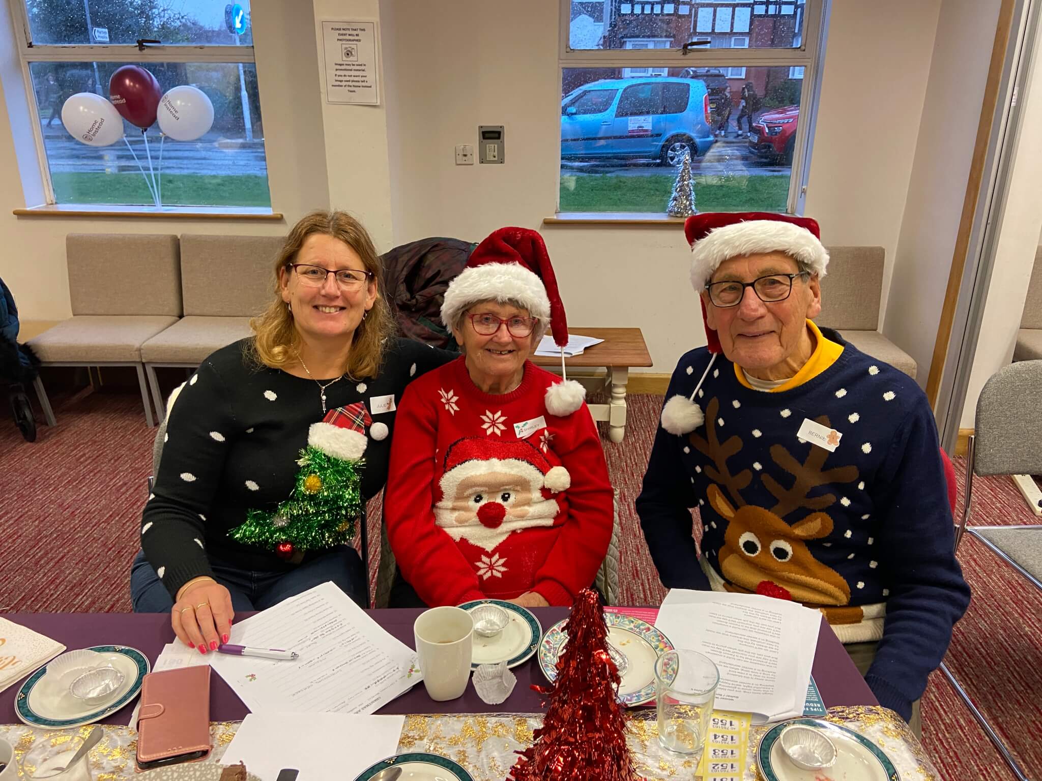 Three people wearing festive sweaters and Santa hats sitting at a table with papers and holiday decorations. - Home Instead