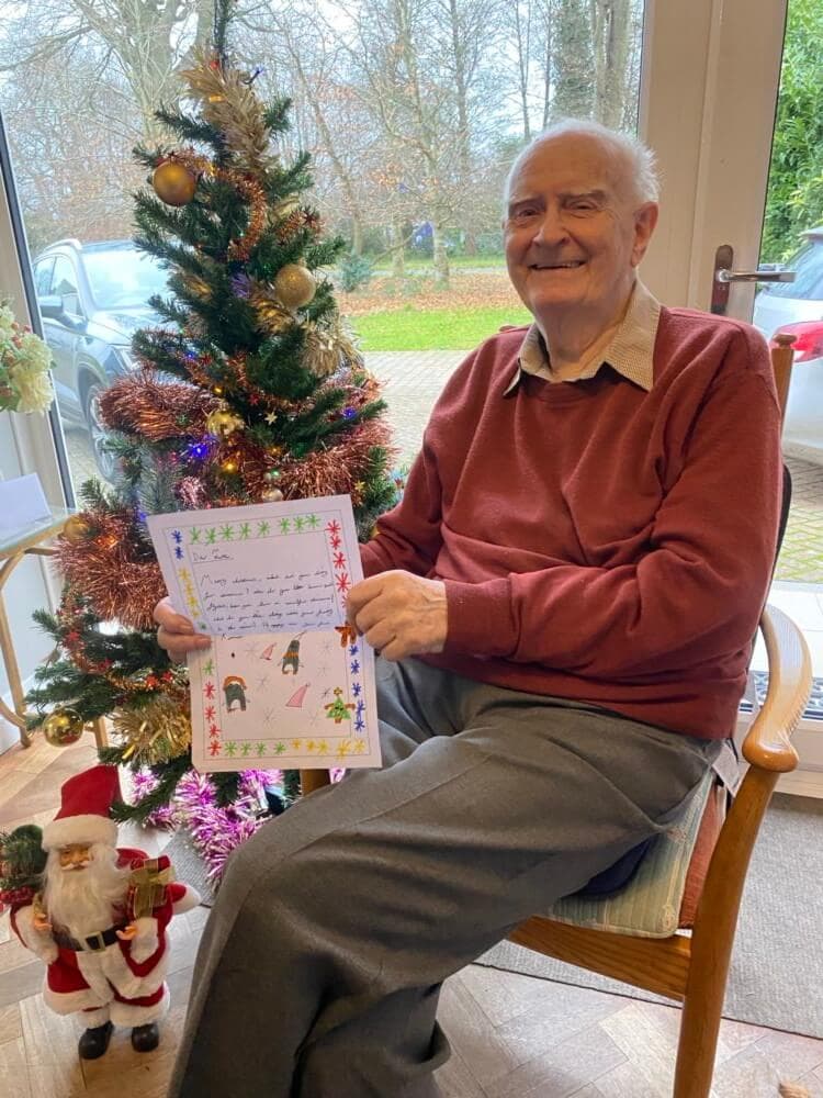 An elderly man sits smiling by a Christmas tree, holding a hand-drawn holiday card. A Santa figurine is at his feet. - Home Instead