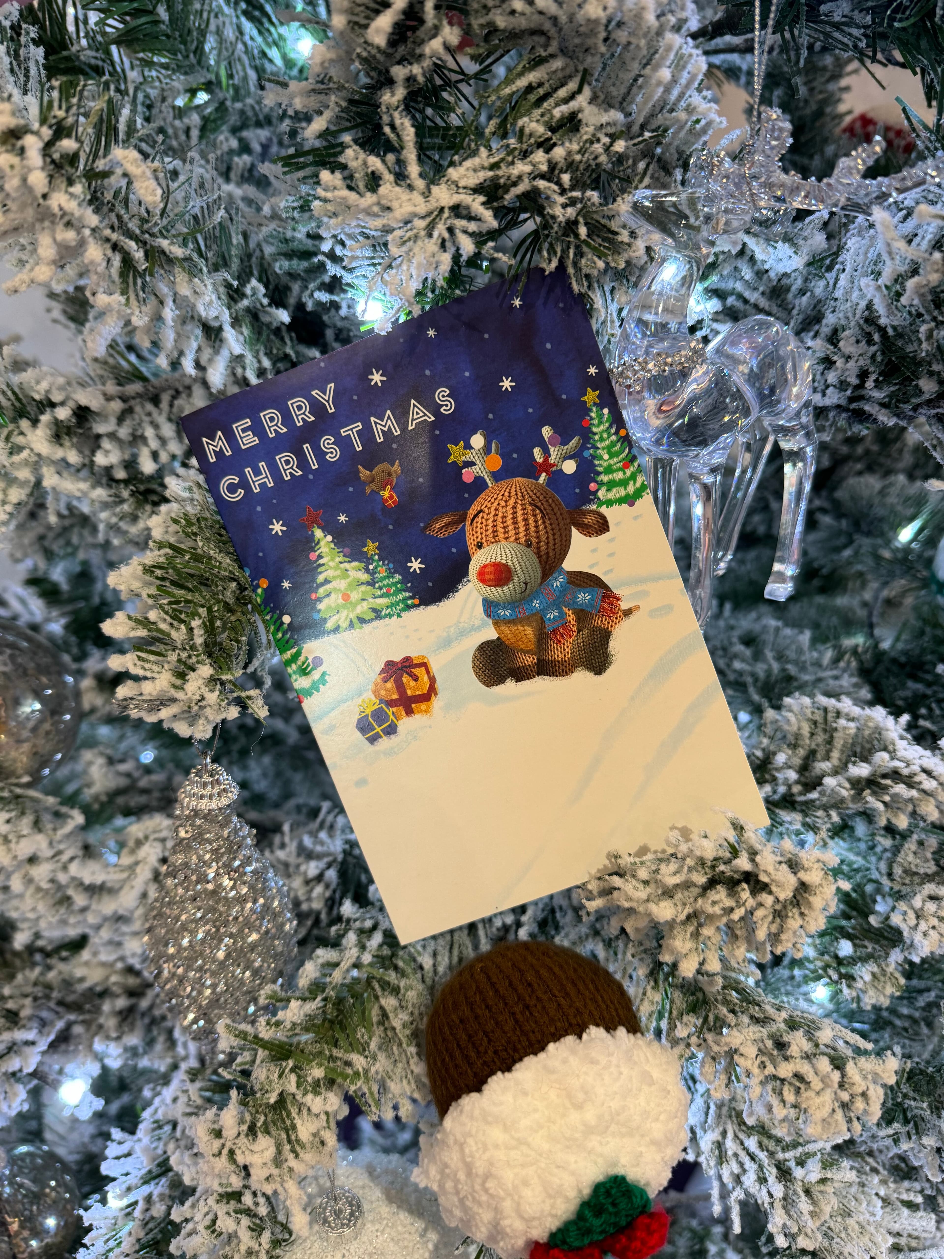 A Christmas card with a reindeer on a snowy scene hangs on a decorated, snowy Christmas tree. - Home Instead