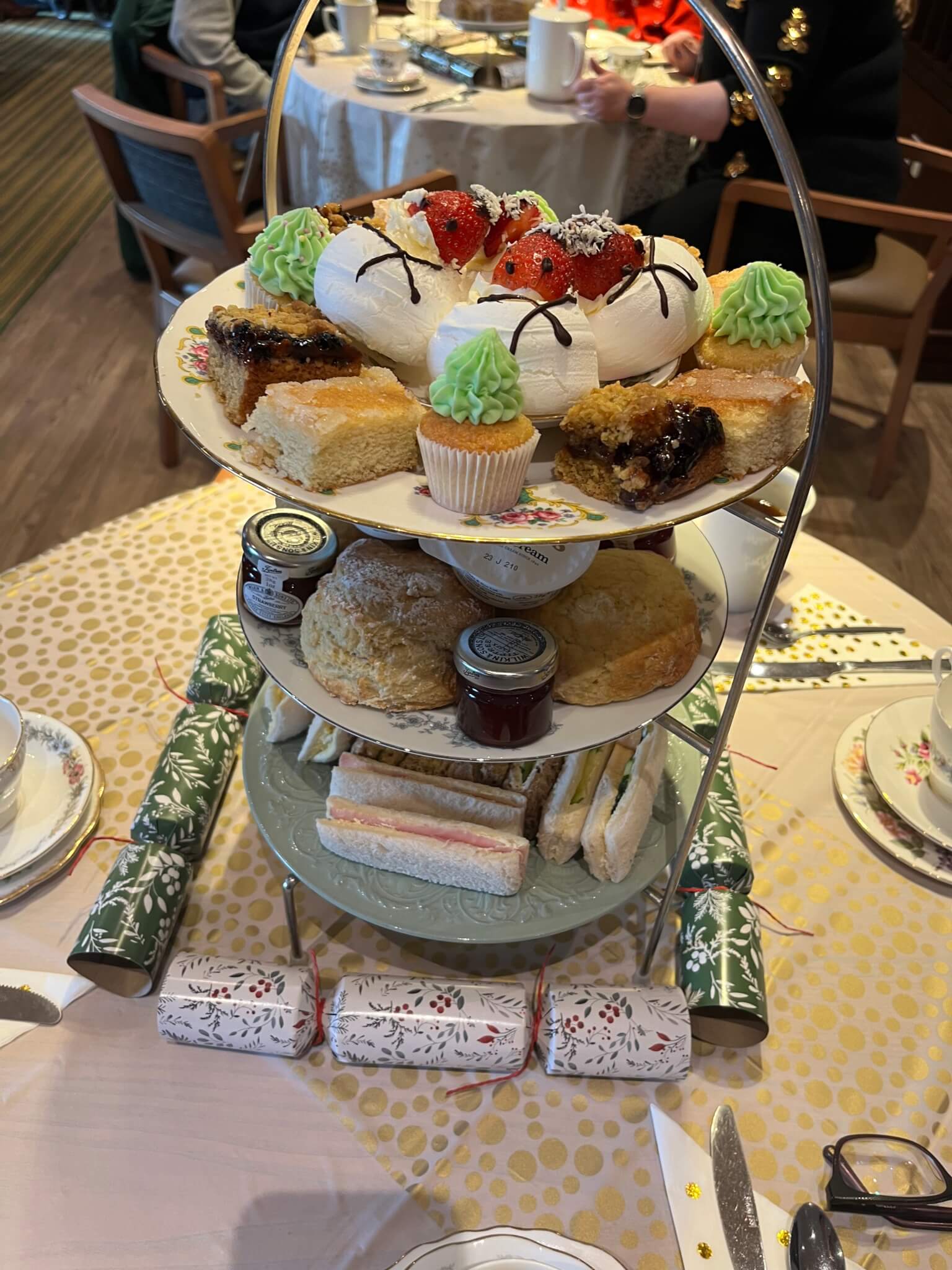 A three-tiered afternoon tea stand with various pastries, sandwiches, scones, and Christmas crackers on a table. - Home Instead