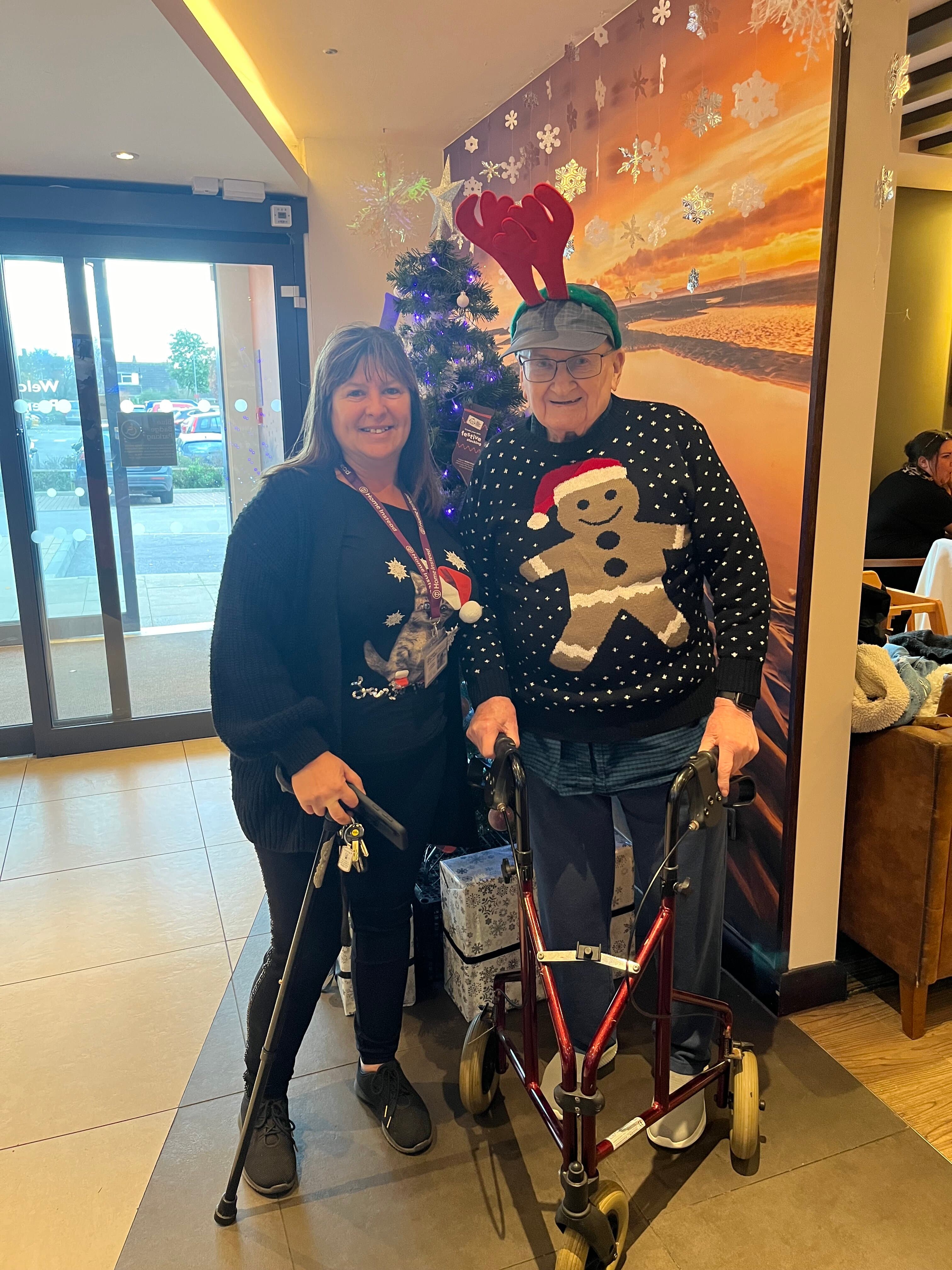 Two people in Christmas sweaters stand next to a festive tree, one uses a walker and wears reindeer antlers. - Home Instead
