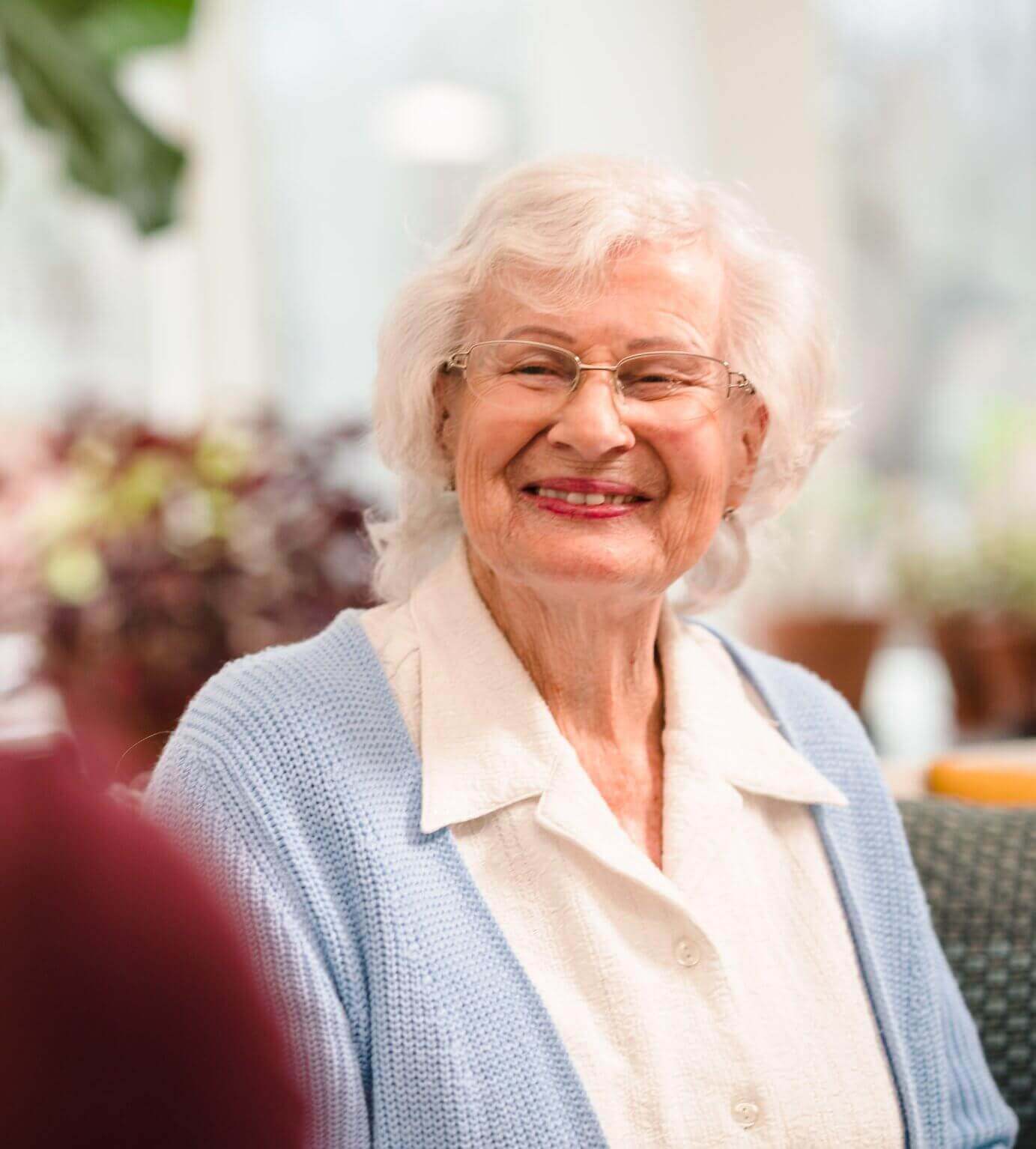 Elderly woman with glasses and white hair, smiling warmly, wearing a light blue cardigan over a white blouse. - Home Instead