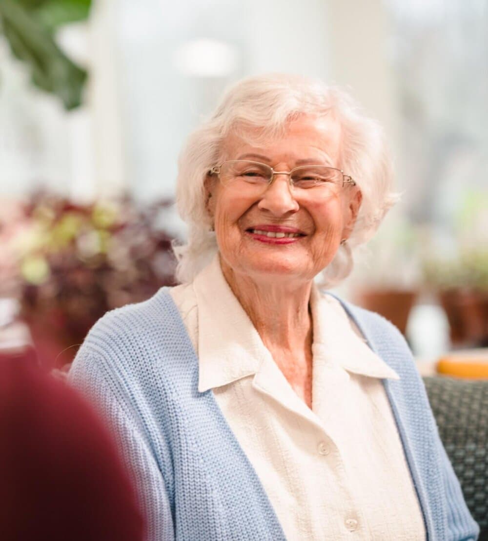 Elderly woman with white hair and glasses smiling, wearing a white blouse and blue cardigan, indoors. - Home Instead