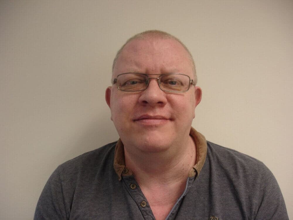 A man with glasses wearing a collared shirt smiles at the camera against a plain background. - Home Instead