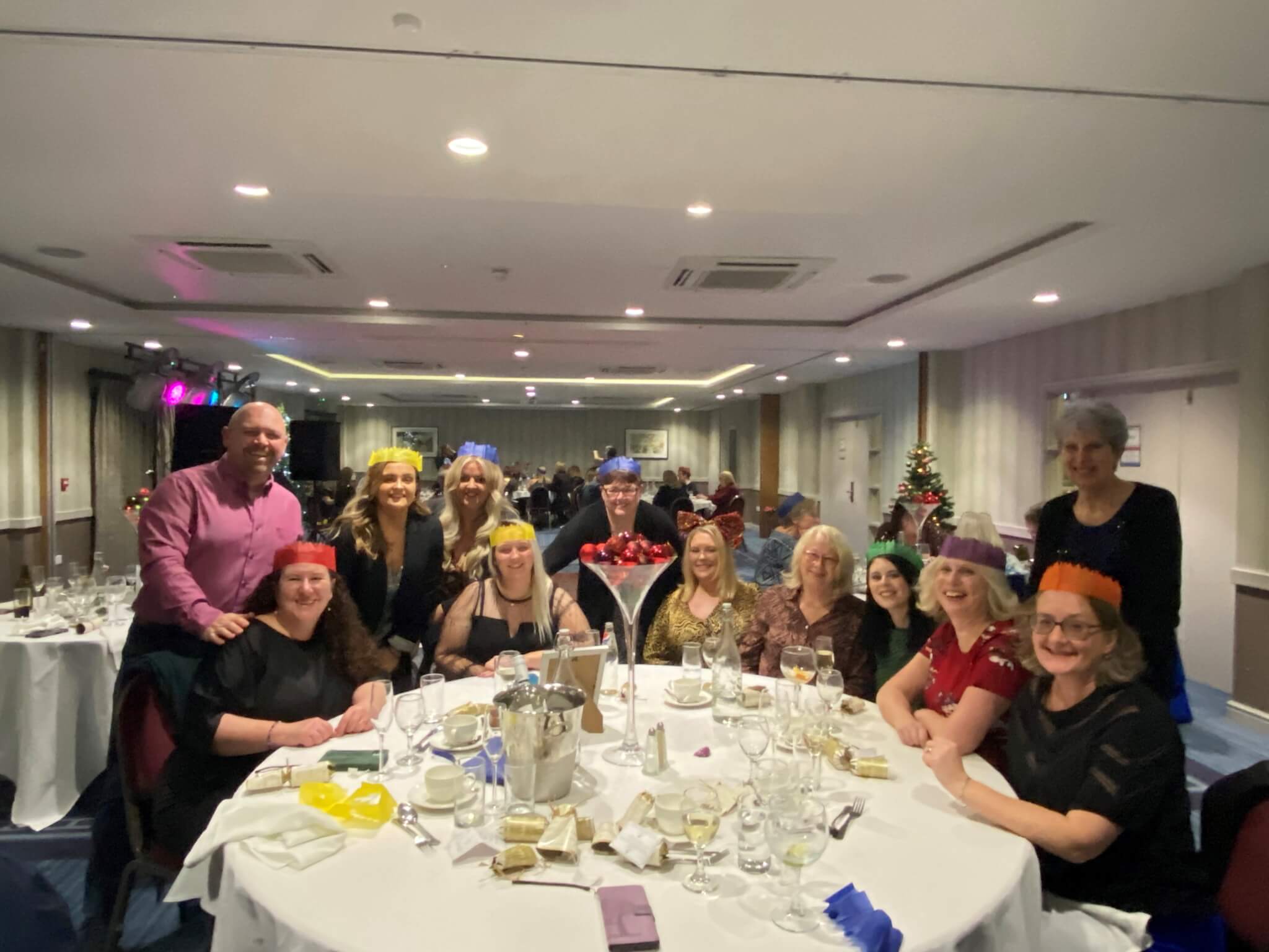 A group of people in party hats around a decorated table at a festive gathering inside a function room. - Home Instead