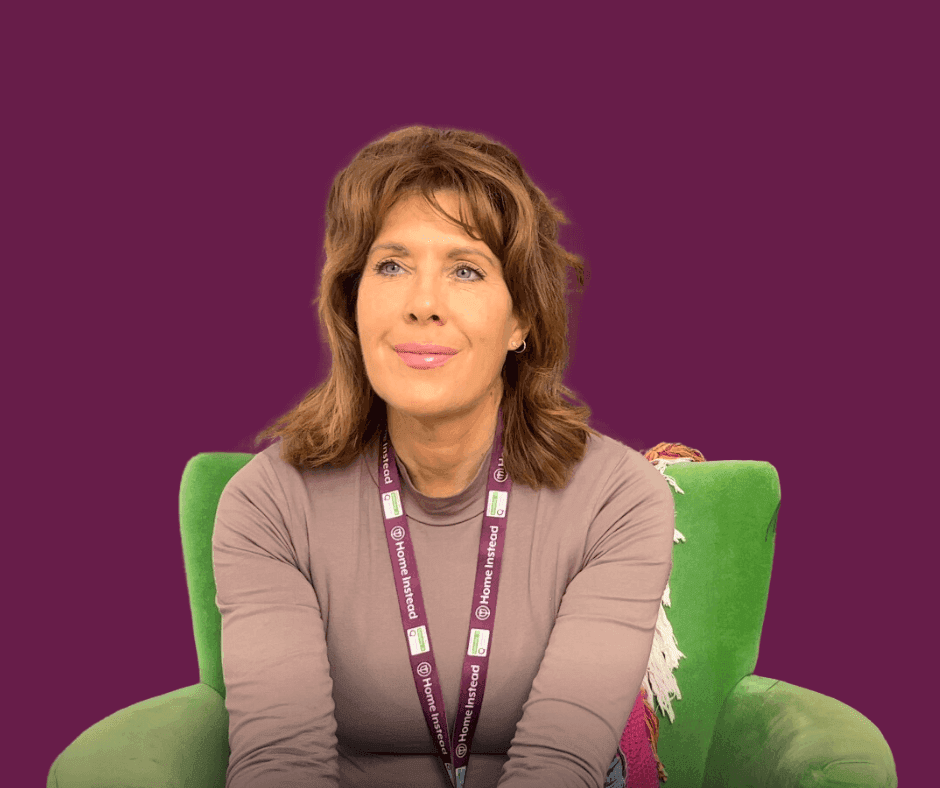 A woman with light brown hair sits on a green chair, against a purple background, wearing a lanyard and a gray top. - Home Instead