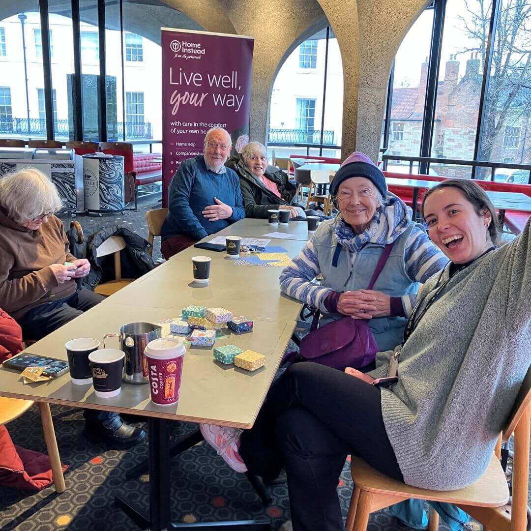 A group of cheerful seniors and a young woman sit around a table with coffee cups at a social event indoors. - Home Instead