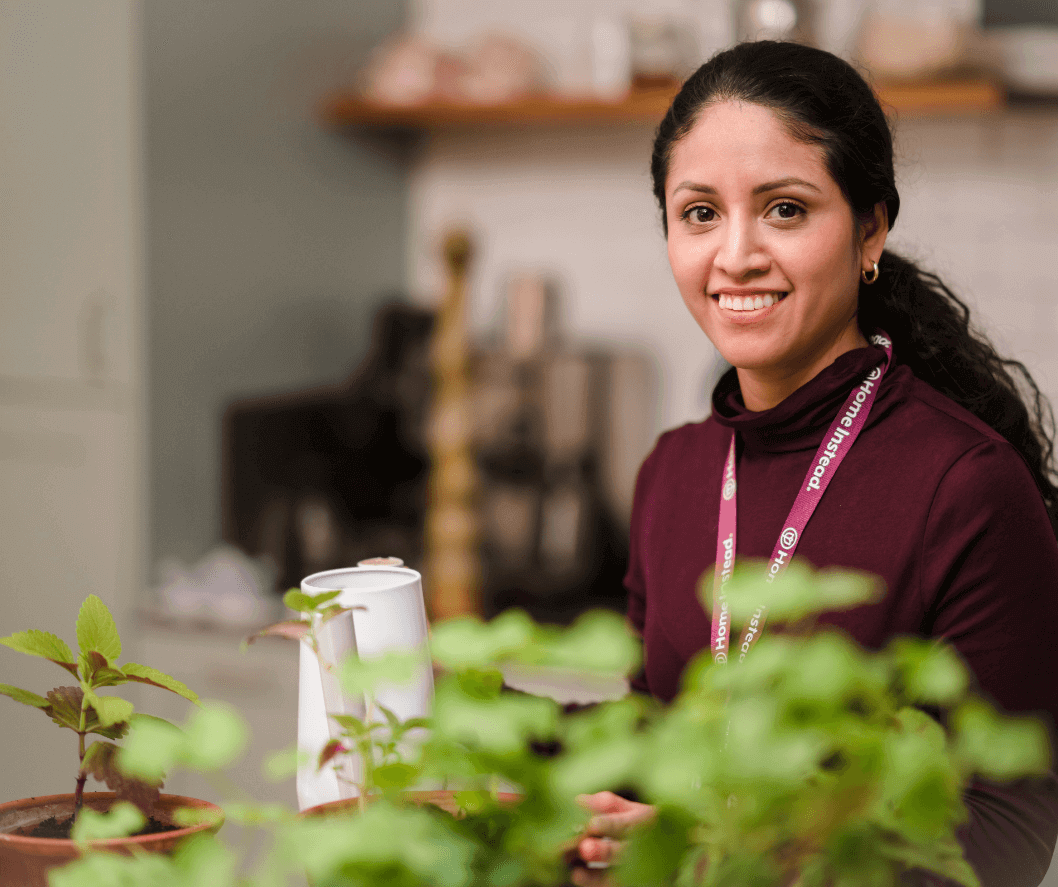 A woman with a lanyard smiles while sitting at a table with small potted plants. Kitchen appliances are in the background. - Home Instead