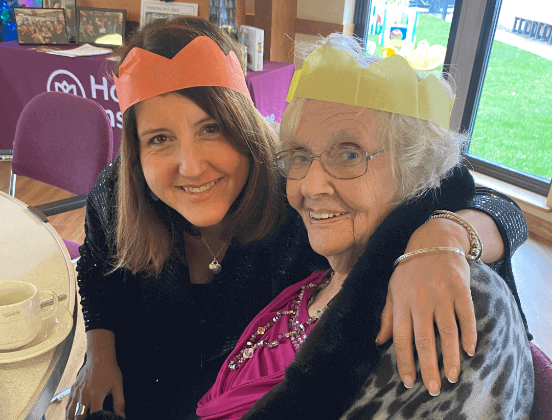 Two women wearing paper crowns and smiling, one older and one younger, sit together at a table. - Home Instead