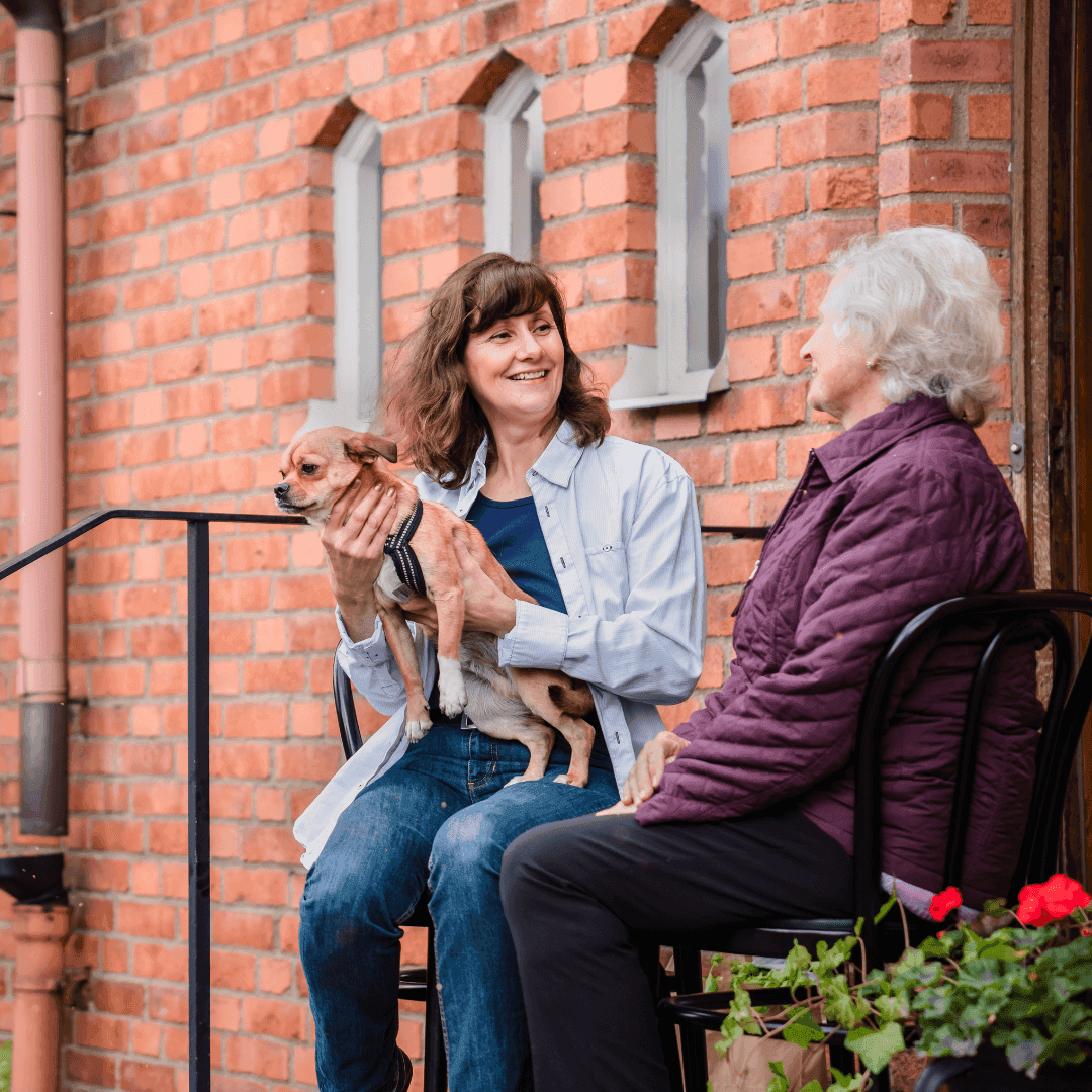Female Live-in Care Professional holding a dog and a female client sat on the steps of a house