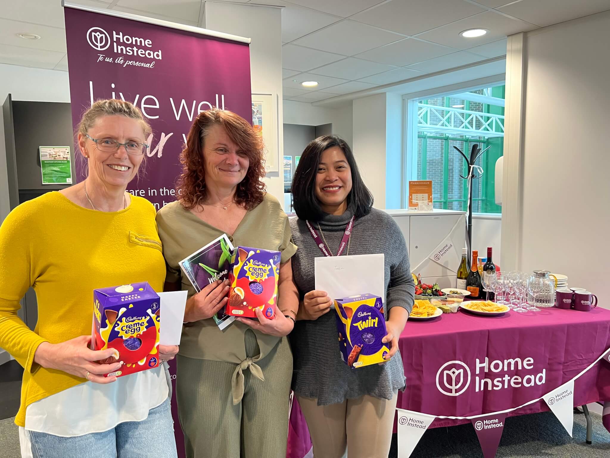 Three women holding Easter eggs, standing in front of a Home Instead table with food and drinks displayed. - Home Instead