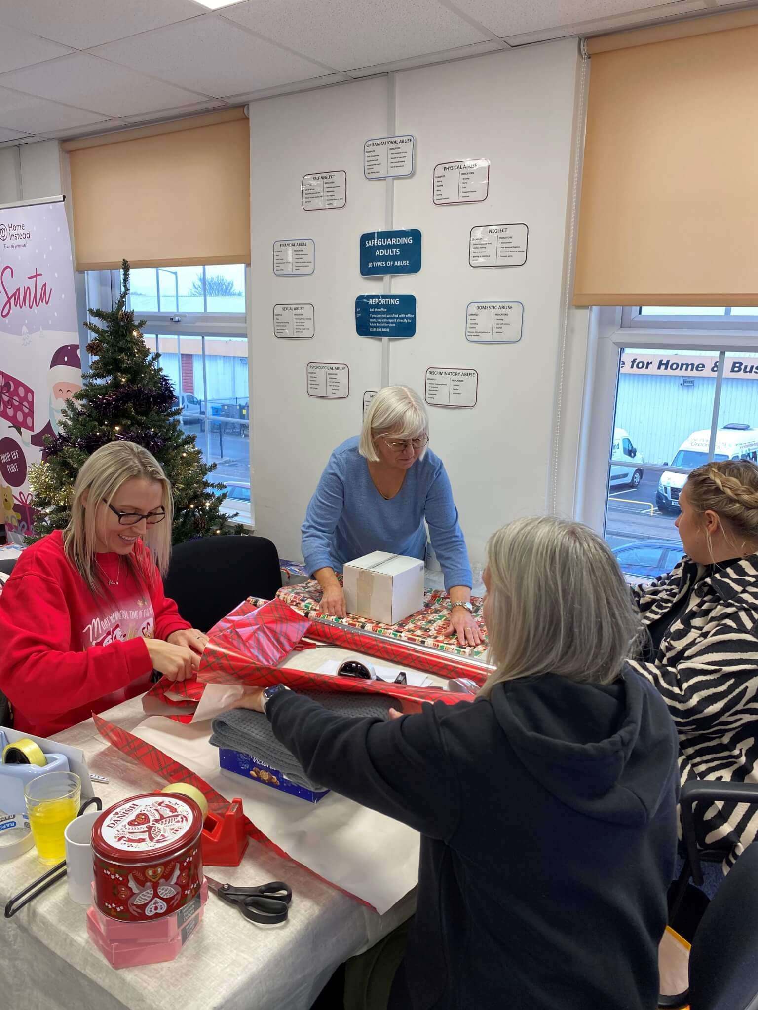 Four women are wrapping gifts at a table in a festive room with a Christmas tree and decorations. - Home Instead
