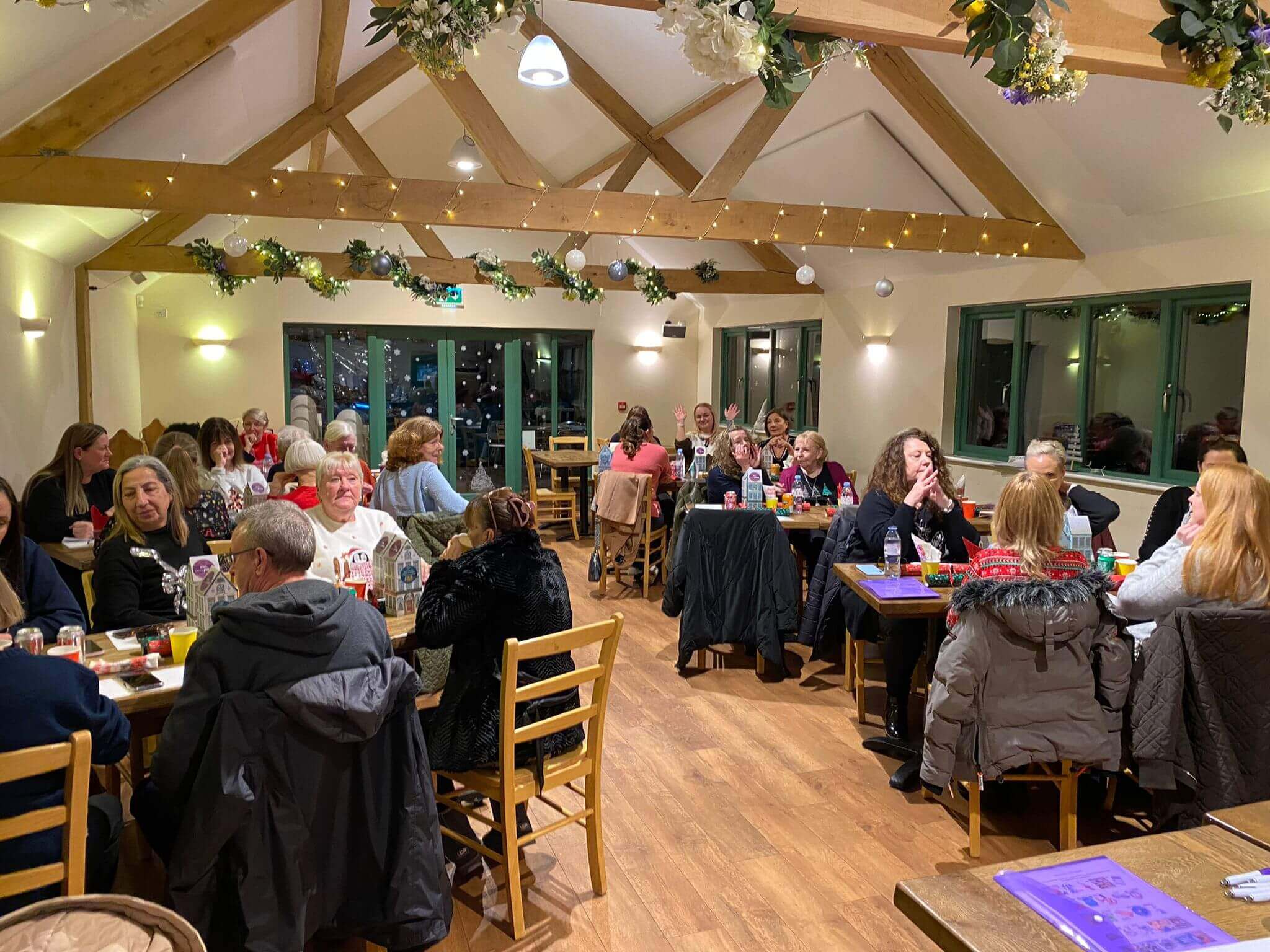 People seated at tables for an indoor gathering in a decorated room with wooden beams and festive lights. - Home Instead