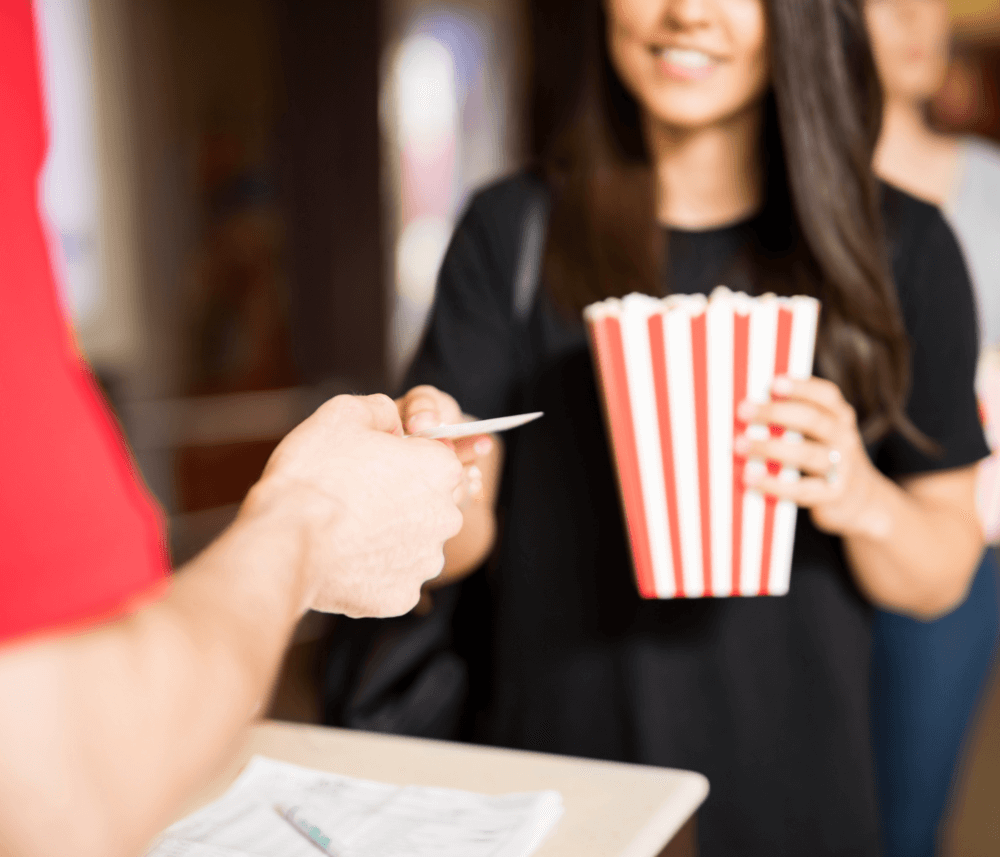 Person handing a ticket to staff while holding a red and white striped popcorn container. - Home Instead